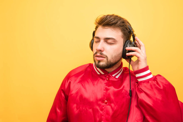 Portré egy férfi a szakálla, fárasztó egy piros kabát, áll a háttérben egy sárga fal, és ő hallgat zenét a fejhallgatót. Az ember szeret zenét hallgatni. Izolált sárga háttér. - Fotó, kép