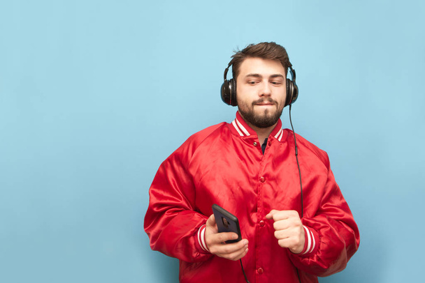 赤いジャケットと青い背景にヘッドフォンで音楽を聴いて手にスマートフォンを着た面白い男。彼の手にスマートフォンで踊るヘッドフォンのひげの男、青い壁. - 写真・画像