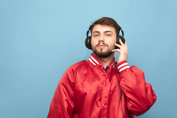 Portret brodatego mężczyzny w słuchawkach i czerwona Kurtka na niebieskim tle, patrząc w aparacie. Dorosły człowiek słucha muzyki w słuchawkach. Izolowane. - Zdjęcie, obraz
