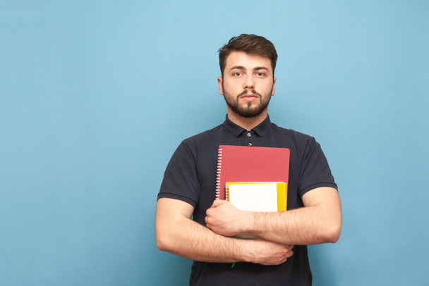 Portret van een student met een baard, het dragen van een donker hemd, staande op een blauwe achtergrond met boeken en notebooks in zijn handen en kijken naar de camera met een ernstig gezicht. Geïsoleerd. - Foto, afbeelding