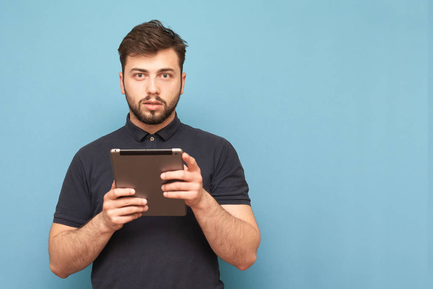 Homme barbu avec une tablette dans les mains se tient sur un fond bleu, regarde dans la caméra, porte un t-shirt sombre. L'homme avec un gadget à la main est isolé sur un fond bleu. Espace de copie
 - Photo, image