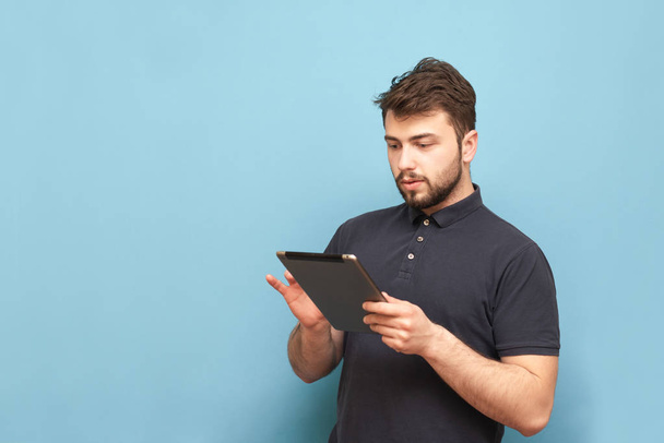 Portret dorosłego skoncentrowanego człowieka korzysta z Internetu na tablecie, ubrany w brodę i ciemny T-shirt. Izolowane. Człowiek biznesowy z tabletem w ręku na niebieskim tle. s - Zdjęcie, obraz