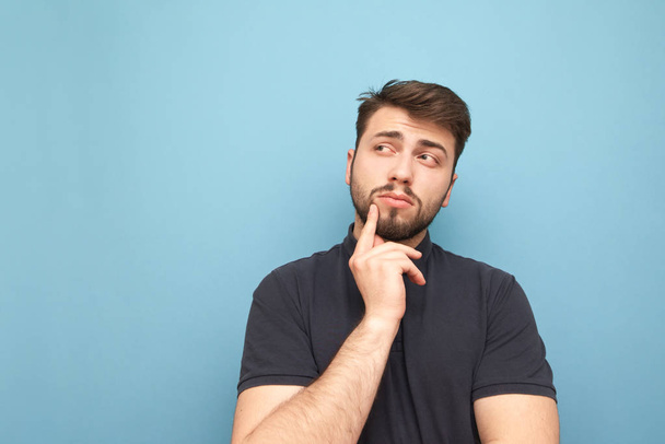 Man with a beard and dressed in a dark T-shirt thinks on a blue background and looks sideways on ��opyspace. Thoughtful adult man is isolated on a blue background. - Foto, Bild