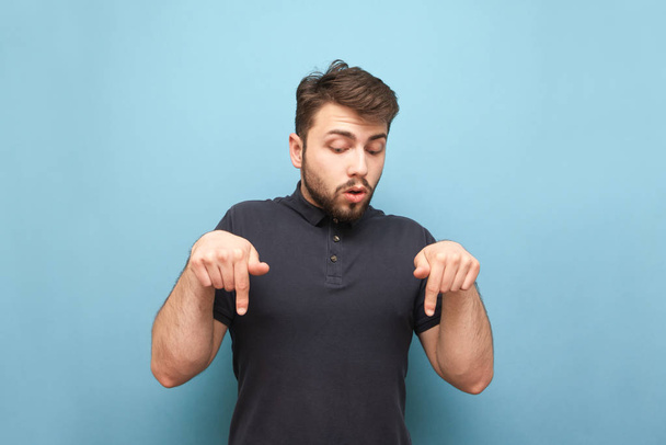 Portrait of a surprised man on a blue background, looking down and showing his fingers. Man with a beard shows hands on copyspace, isolated - Foto, immagini