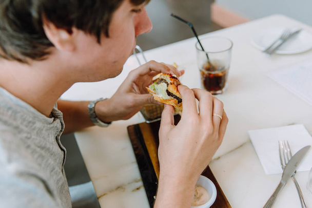 young man in casual clothes eatig a delicious burger in a cafe  - Фото, изображение