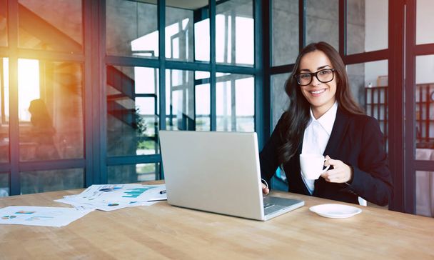 Portrait of smiling pretty young business woman in glasses and full suit sitting on workplace at office - Fotoğraf, Görsel