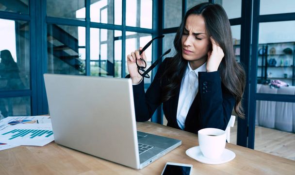 Κουρασμένη γυναίκα επιχείρηση σε πλήρες κοστούμι με πονοκέφαλο ενώ εργάζεται για το laptop της στο σύγχρονο γραφείο κρατά το κεφάλι της. - Φωτογραφία, εικόνα