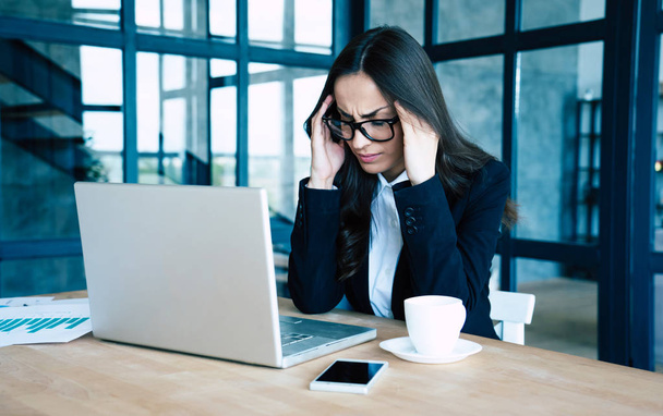 Κουρασμένη γυναίκα επιχείρηση σε πλήρες κοστούμι με πονοκέφαλο ενώ εργάζεται για το laptop της στο σύγχρονο γραφείο κρατά το κεφάλι της. - Φωτογραφία, εικόνα