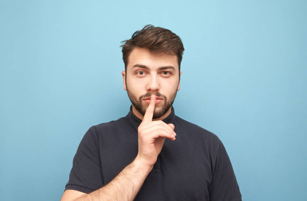 Man in a dark T-shirt on a blue background holds his finger around his mouth and looks thoughtfully at the camera.Focused man is isolated on a yellow background, a portrait.Man shows a sign of silence - Φωτογραφία, εικόνα