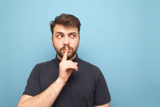 Portrait of an adult man with a beard thoughtfully looking sideways to a blank place and showing a sign of silence holding a finger near his mouth on a blue background. Isolated. - Foto, Bild