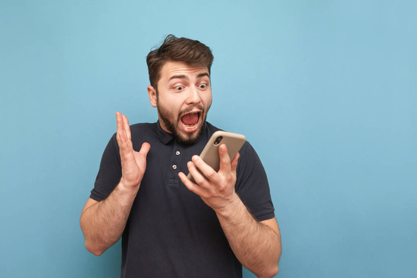Portrait of a crazy bearded man with a smartphone in his hands, looking at the screen and screaming on a blue background. Shocked man uses a smartphone and rejoices. Isolated. - Fotoğraf, Görsel