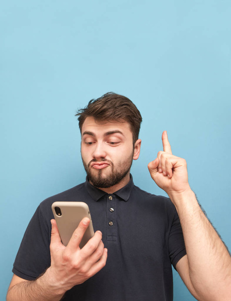 Emotionaler Mann mit Bart hält ein Smartphone in der Hand und zeigt den Finger in eine Leerstelle. Überraschter Erwachsener in dunklem T-Shirt benutzt Smartphone und zeigt Finger auf Copyspace. - Foto, Bild