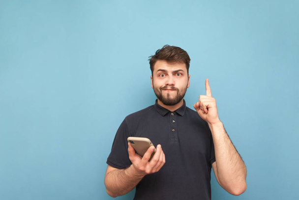 Portrait of a funny man with a beard and a phone in his hand on a blue background, looking into the camera and making a funny face. Confused man with a smartphone shows an empty space. Isolated. - Foto, Bild