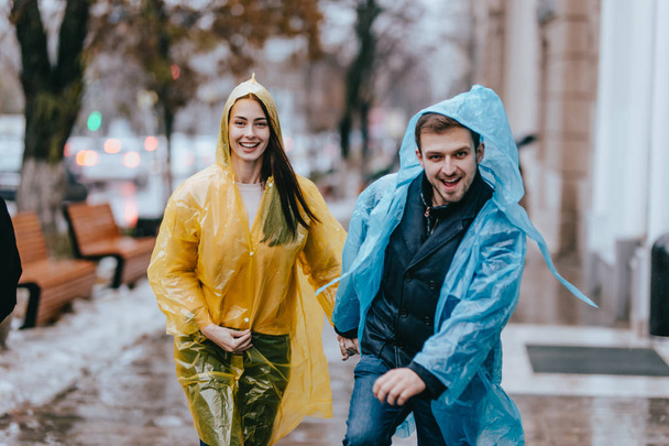 Funny and loving guy and girl in yellow and blue raincoats are running in the rain outside. - Fotoğraf, Görsel