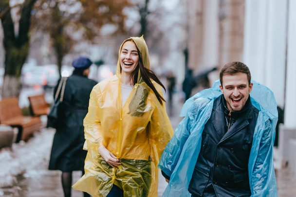 Funny and loving guy and girl in yellow and blue raincoats are running in the rain outside. - Фото, изображение