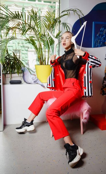 Fashion young girl blogger dressed in stylish striped jacket and red trousers poses sitting on the stool with pink fur in the room on the background with plant - Photo, image
