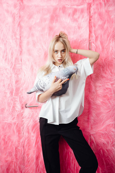 Young girl blogger dressed in stylish white shirt and black trousers poses with with a soft toy in the shape of a pigeon on the background of pink fur wall - Photo, Image