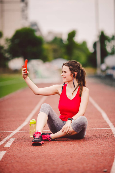 Νεαρή αθλήτρια στο στάδιο, αθλητική ζωή, κάθεται στην πίστα, βγάζει selfie φωτογραφία σε ένα smartphone, χαμογελώντας χαρούμενη στο ακουστικό των αυτιών της, κοντά σε φιάλη φιάλης με νερό - Φωτογραφία, εικόνα