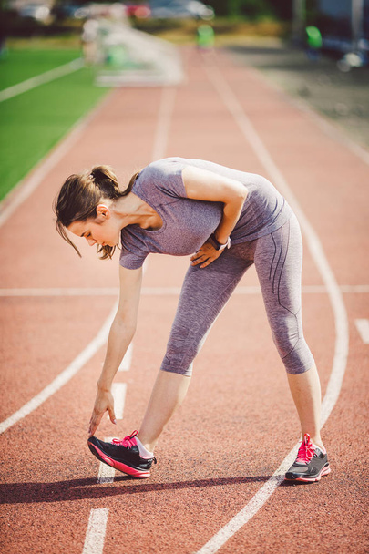Beautiful young caucasian woman with long hair in tail and big breasts doing exercises, warming up and warming up muscles before training in running stadium, red rubber track in summer on a sunny day - Photo, Image