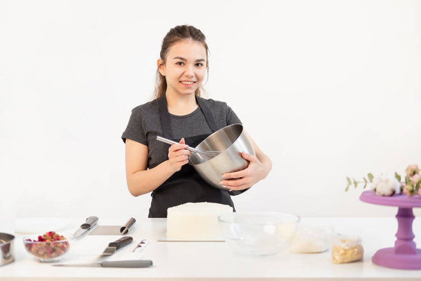 Attractive brunette girl pastry chef in apron whips cream cake in a metal bowl. - 写真・画像