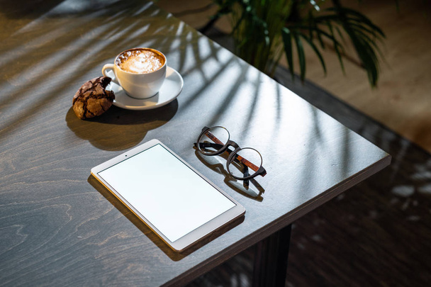 Nahaufnahme von Kaffeetasse, Keksen, Gläsern und Block mit leerem Bildschirm auf dem Tisch. - Foto, Bild