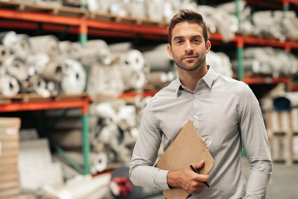 Portrait of a manager holding a clipboard while standing on a carpet warehouse floor with inventory on shelves in the background  - Foto, imagen