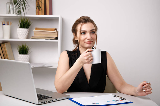 Woman in office drinking coffee at her workplace near the computer. - 写真・画像