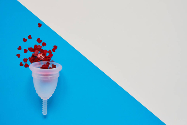 Bílý menstruální pohár s červenými třpyty na modrém pozadí. Alternativní produkt ženské hygieny v daném období. Zdravotní koncepce žen. Kopírovat místo - Fotografie, Obrázek