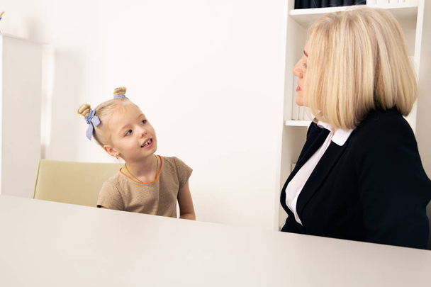 Female psychologist with little kid patient sitting indoor at the desk and talking. Child problem concept. - Photo, Image