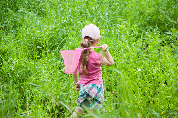 A little girl in a pink T-shirt stands with a net in her hands. Catching butterflies. On a green meadow in summer. Green grass. - Fotoğraf, Görsel