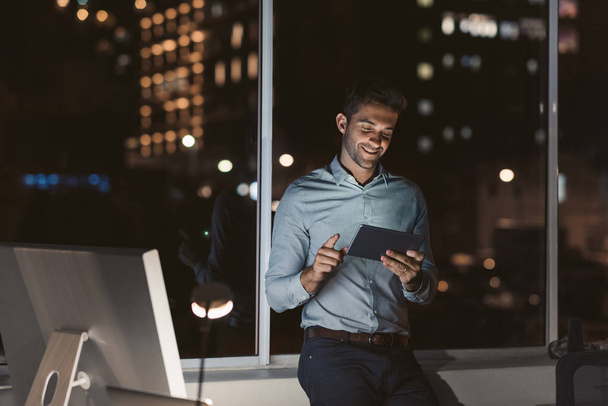 Smiling young businessman standing alone in an office working with a digital tablet late at night with city lights in the background - Zdjęcie, obraz