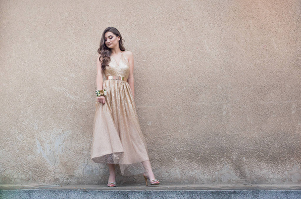 Beautiful teen girl in glamorous golden dress standing by the wall. - 写真・画像