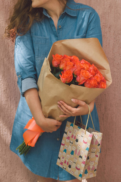 young woman in denim dress with bouquet of roses and gift paper bag - Фото, изображение