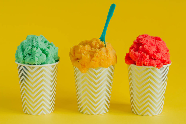 green,yellow and red fruit ice cream or frozen yogurt in stripped    cups  with a spoon on a yellow background - Foto, Imagen