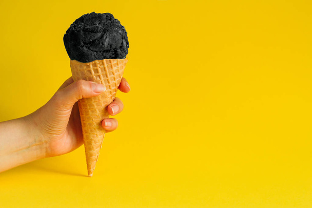 female hand holding waffle cone ice cream with black coal isolated on a vibrant yellow background, copy space - Fotografie, Obrázek