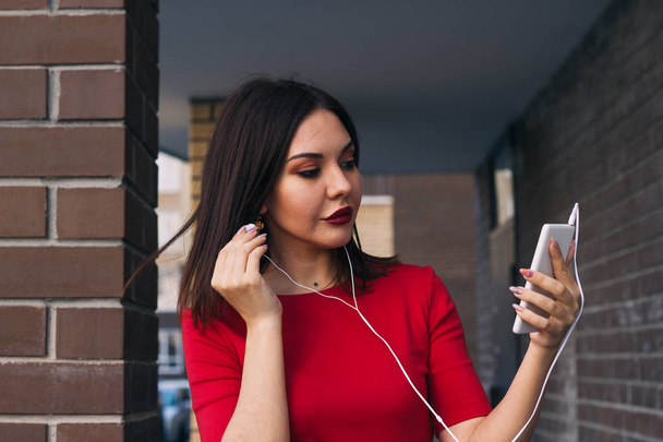 beautiful young woman with red lipstick in red dress uses phone and earphones - Foto, immagini