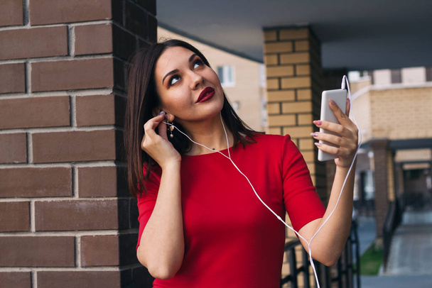 beautiful young woman with red lipstick in red dress uses phone and earphones - Fotó, kép