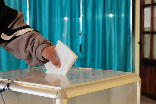 V dotazní stanici. Close-up. Mužská ruka umístí hlasovací papír do průhledné krabice. Modrý pozadí. Banner. Celostátní hlasování, volby. - Fotografie, Obrázek