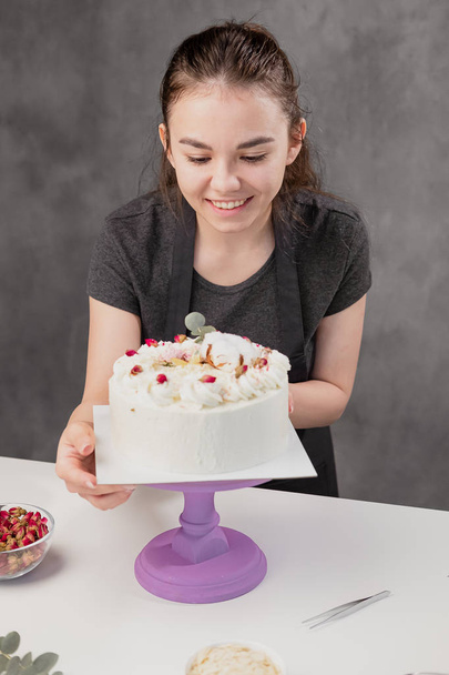 Attractive young woman brunette confectioner presents a white cake with small red flowers of a food rose. - Foto, imagen