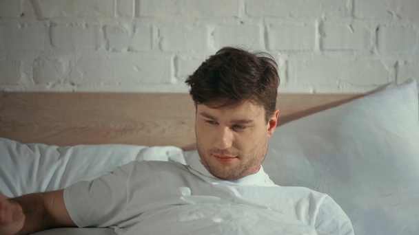 człowiek w białym t-shirt spania w łóżku, budząc się i patrząc na aparat z uśmiechem - Materiał filmowy, wideo