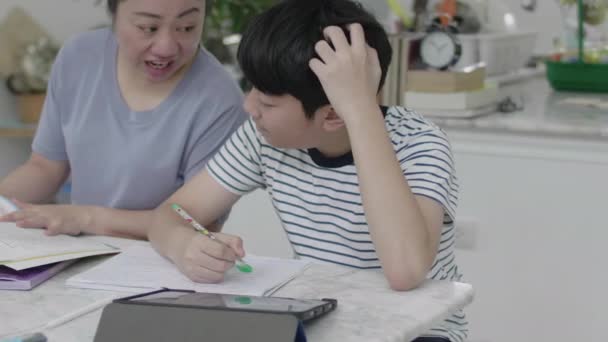 Mother helping her son to do his homework at home. Beautiful mother reading textbook and talking with son while doing homework together at home. - Footage, Video