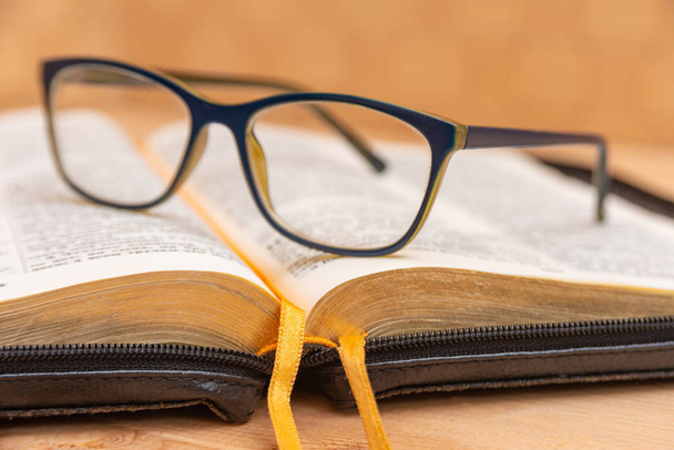 Okulary dla wzroku leżą na Biblii, Biblia w skórzanej czarnej okładce z kartami leży na stole. - Zdjęcie, obraz