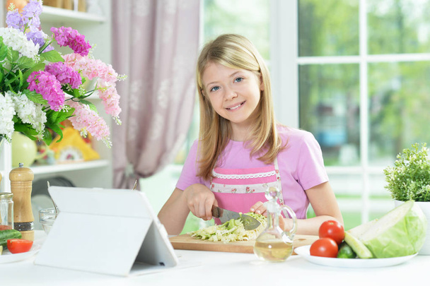 Cute little girl preparing fresh salad on kitchen table with tablet at home - Zdjęcie, obraz