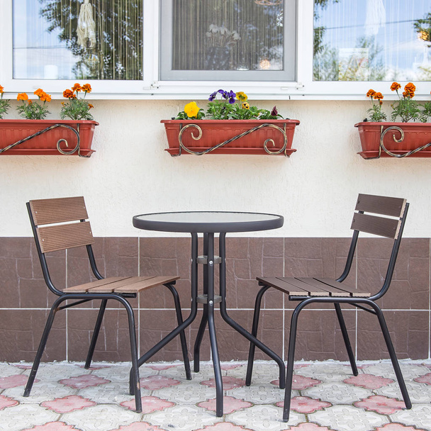 Стеклянные и металлические столы и стулья на летней террасе кафе у окна с горшками цветов
 - Фото, изображение