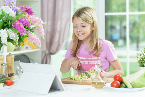 Cute little girl preparing fresh salad on kitchen table with tablet at home - Фото, изображение