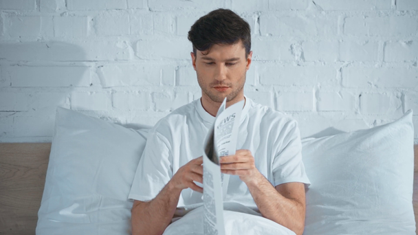 człowiek w białym t-shirt siedzi na łóżku, czytanie gazet i toczenia strony - Materiał filmowy, wideo