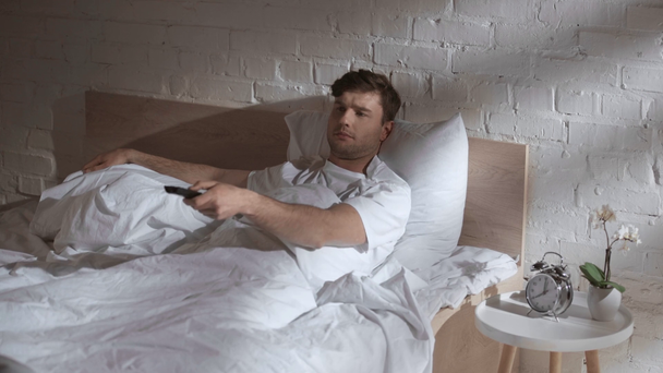 man lying under blanket on bed, holding remote controller, changing tv program and putting remote controller back - Felvétel, videó