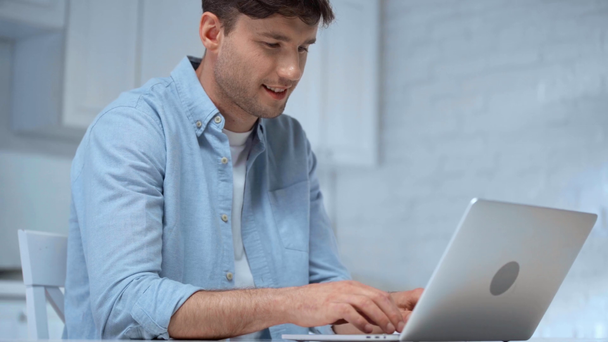freelancer riendo en camisa azul sentado en la cocina y escribiendo en el teclado del ordenador portátil
 - Metraje, vídeo