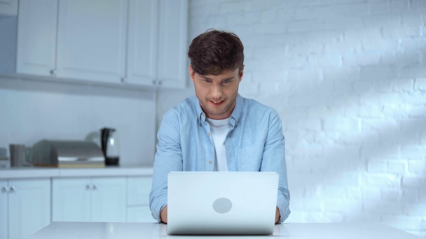freelance excité en bleu chemise ouverture ordinateur portable et dactylographier sur le clavier avec sourire dans la cuisine
 - Séquence, vidéo