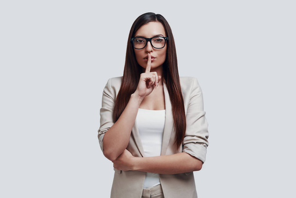 Can you keep a secret? Attractive young woman looking at camera and keeping finger on lips while standing against grey background - Photo, Image